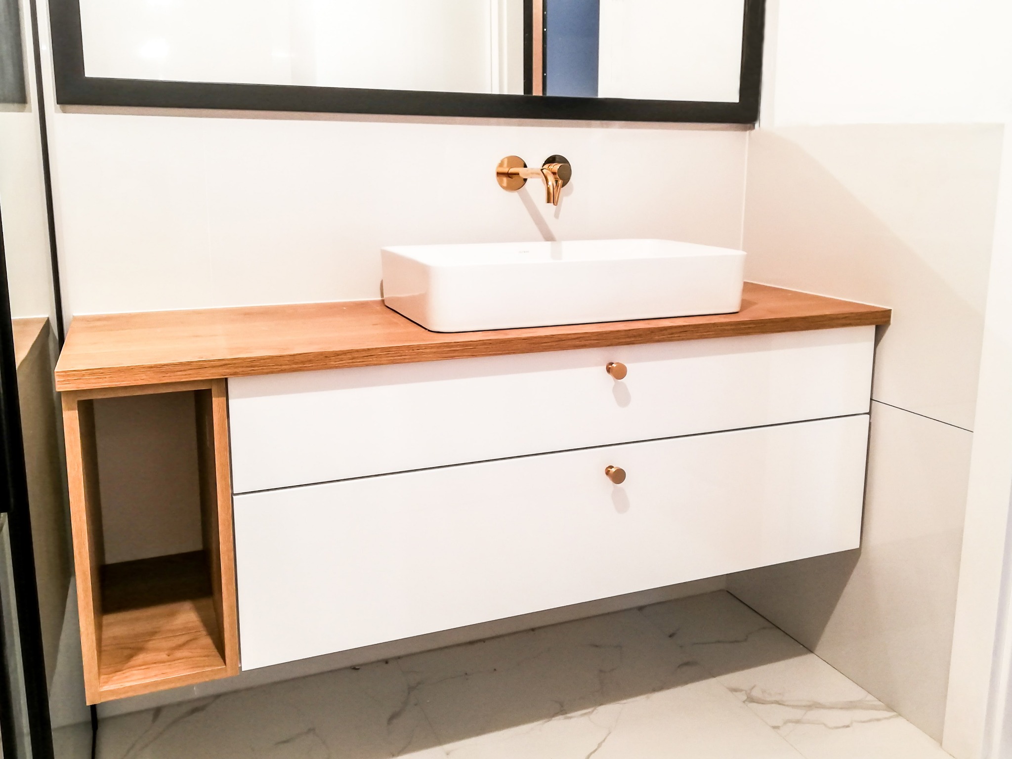 Nowoczesna łazienka z białą, prostokątną umywalką nablatową i złotą baterią ścienną, umieszczona na szafce z drewnianym blatem i białymi frontami szuflad z okrągłymi, złotymi uchwytami. Po lewej...