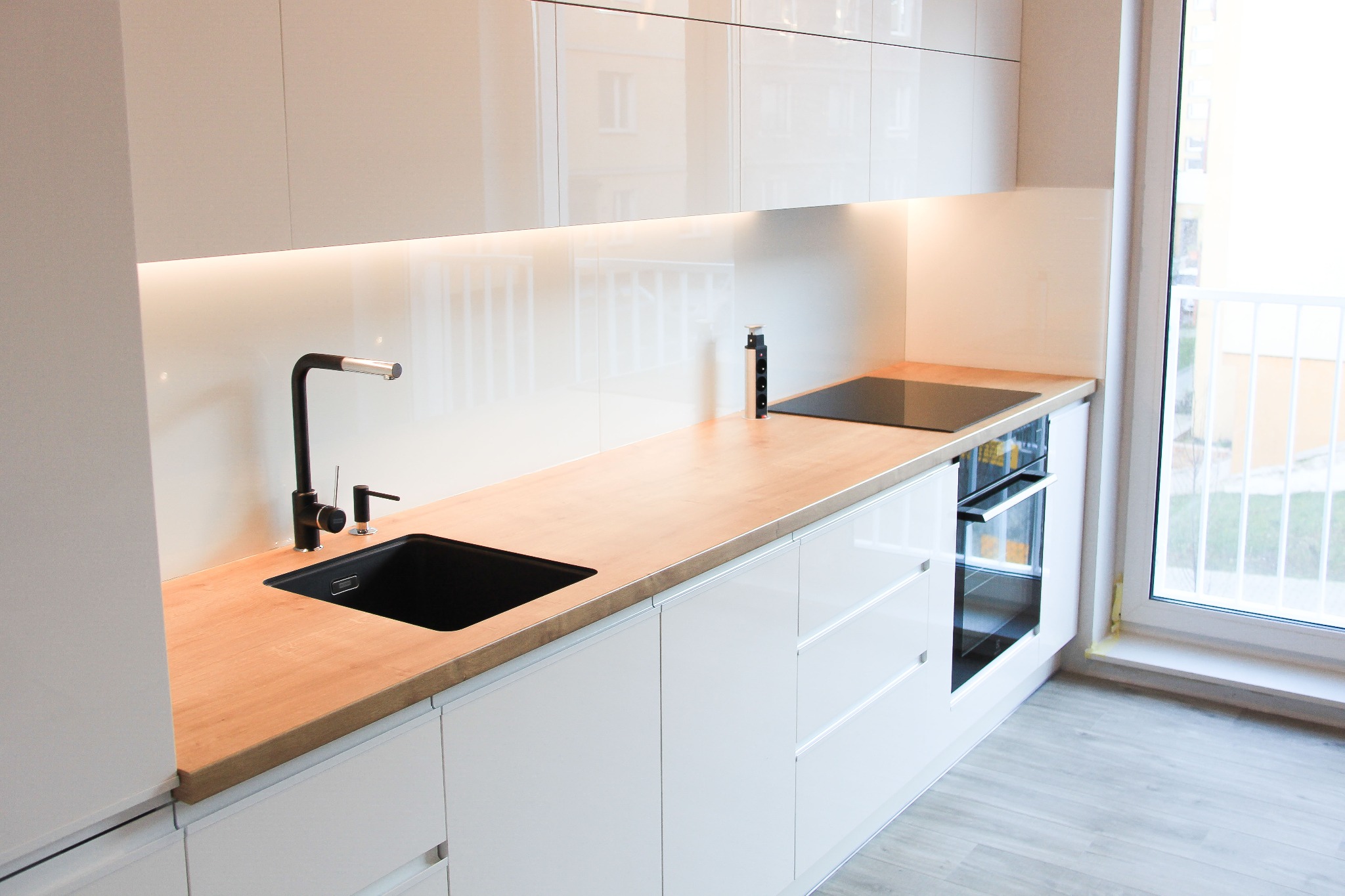 Nowoczesna kuchnia z białymi, lakierowanymi frontami szafek, drewnianym blatem, czarnym zlewem i baterią, płyta indukcyjna oraz podświetlenie LED pod szafkami górnymi.