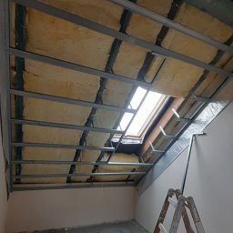 Tomasz skalka - Ocieplenie poddasza z widoczną wełną mineralną, metalową konstrukcją stelaża i oknem dachowym, drabina oparta o ścianę, fragment instalacji wentylacyjnej z czerwonym zakończeniem.