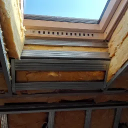 Montaż izolacji z wełny mineralnej w przestrzeni między krokwiami poddasza, widoczne metalowe profile i fragment okna dachowego z widokiem na błękitne niebo.