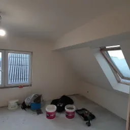 Poddasze po malowaniu, z widocznymi narzędziami malarskimi na podłodze, oknem dachowym i standardowym oknem z widokiem na zimowy krajobraz za oknem.