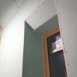 Tomasz skalka - Montaż zielonej płyty gipsowo-kartonowej wokół ościeżnicy okiennej, widoczne wkręty, fragment instalacji elektrycznej pod sufitem.