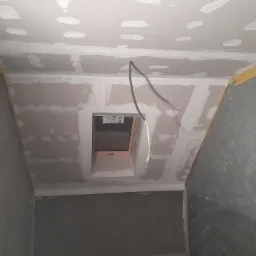 Poddasze w trakcie prac wykończeniowych: płyty karton-gips na skosach i suficie, widoczne łączenia szpachlowane, okno dachowe z drewnianą ramą, wystający kabel elektryczny. Żółta taśma malarska...