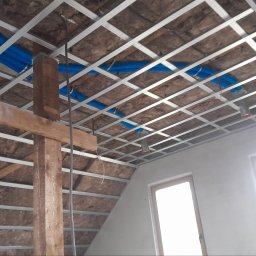 Tomasz skalka - Szkielet z profili metalowych podwieszanego sufitu na poddaszu, z widoczną izolacją z wełny mineralnej i niebieskimi rurami instalacji, drewniany słup podtrzymujący konstrukcję.