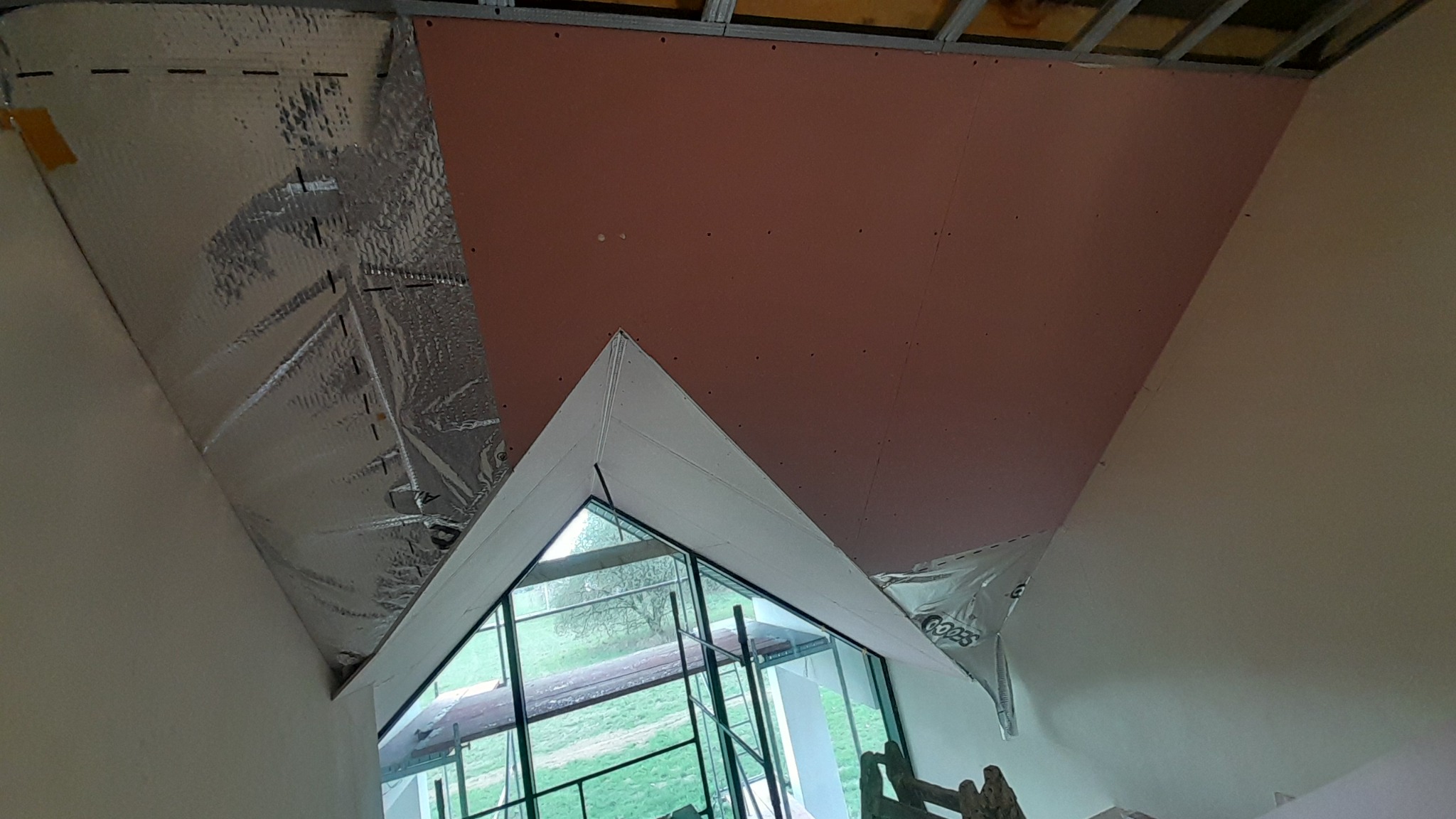 Skośne poddasze w trakcie izolacji: widoczna folia termoizolacyjna i płyta gipsowo-kartonowa na konstrukcji metalowej, okno dachowe w kształcie trójkąta.