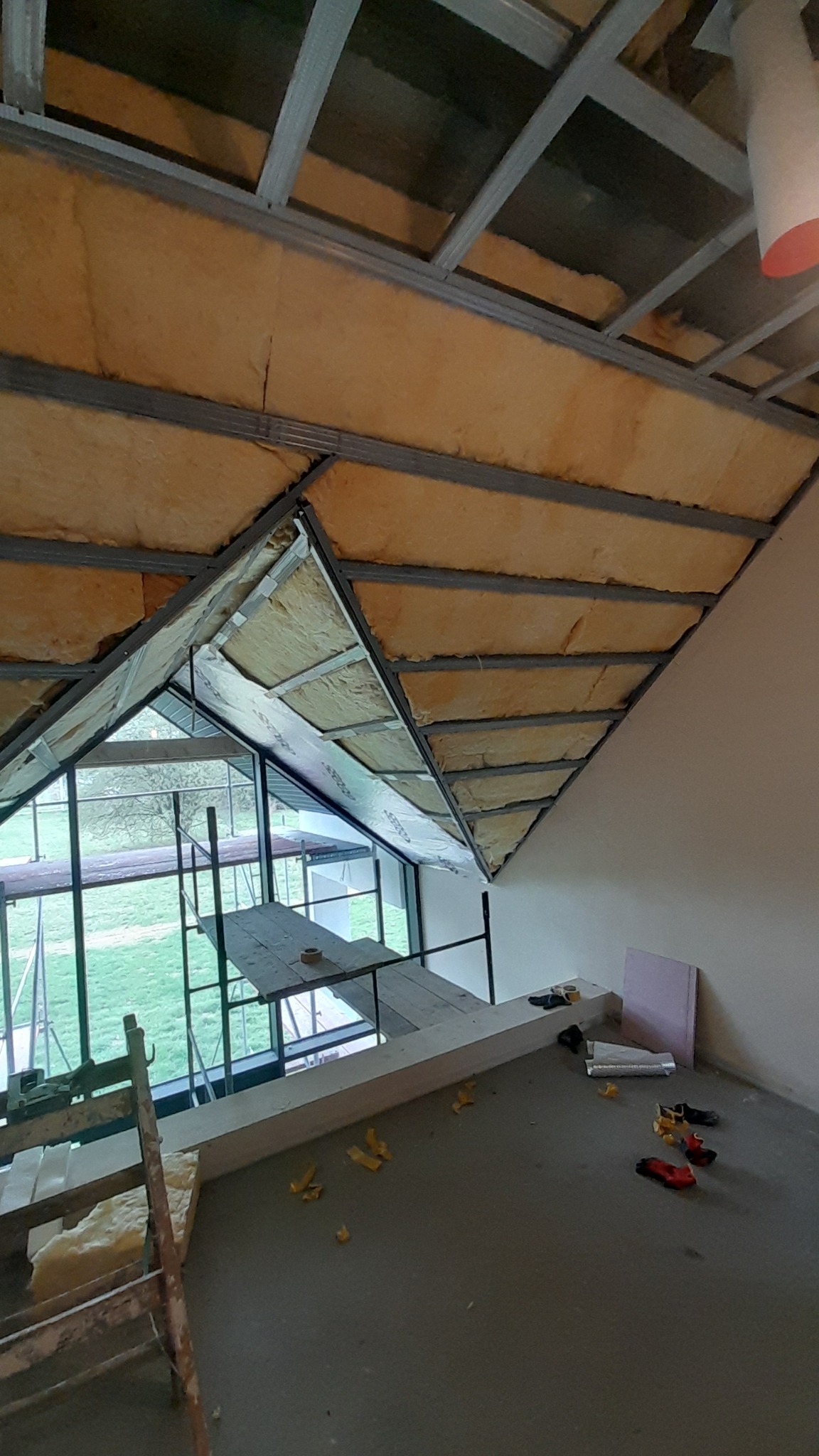 Skośny dach poddasza w Myślenicach w trakcie ocieplania, widoczne metalowe profile, wełna mineralna i folia paroizolacyjna, częściowo zamontowane, z widokiem na zewnątrz przez okno dachowe...