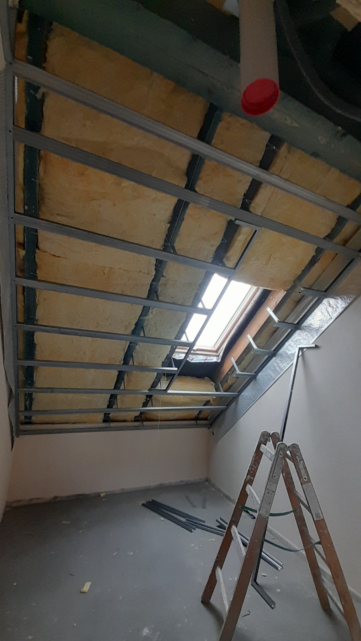Ocieplenie poddasza z widoczną wełną mineralną, metalową konstrukcją stelaża i oknem dachowym, drabina oparta o ścianę, fragment instalacji wentylacyjnej z czerwonym zakończeniem.