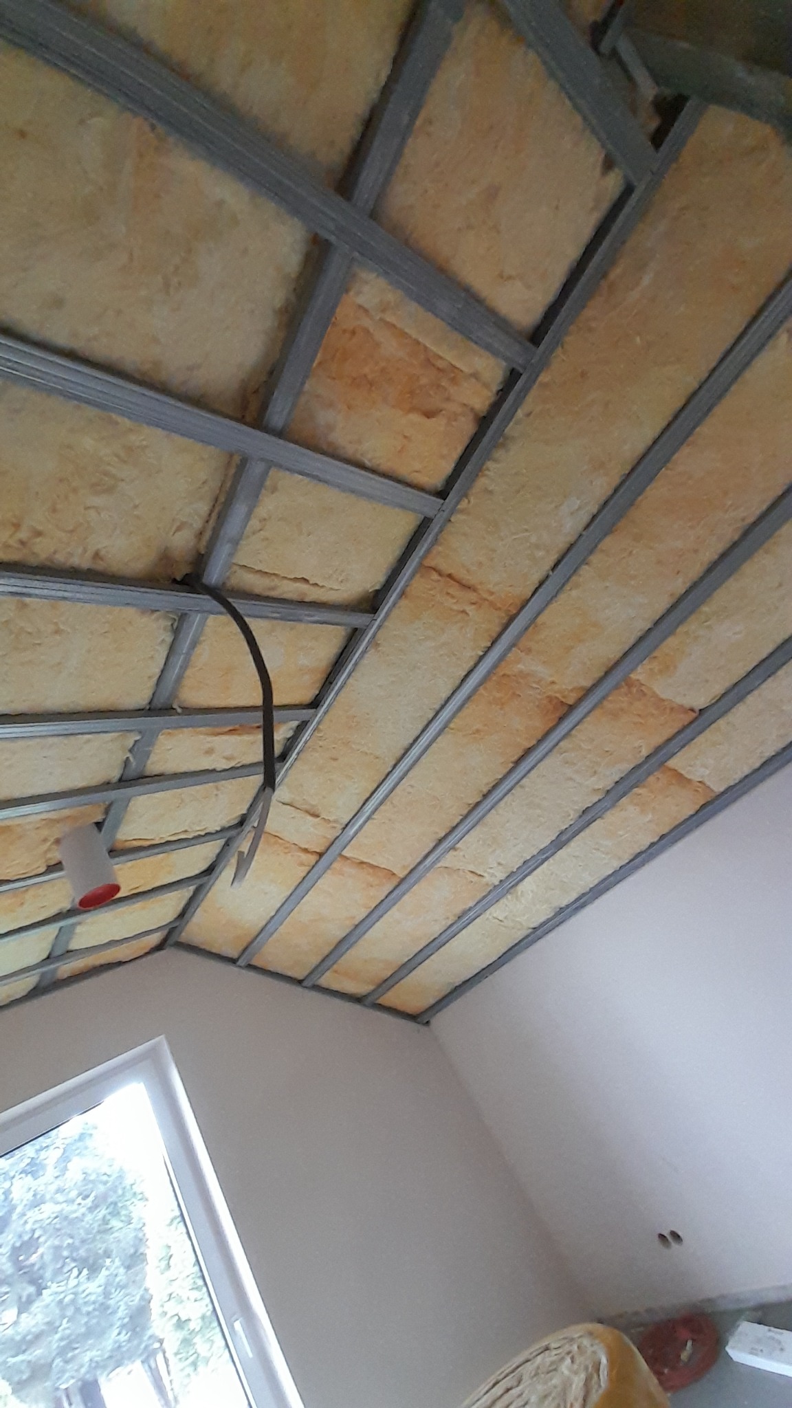 Skośny sufit poddasza z widoczną konstrukcją metalową i izolacją z wełny mineralnej, widok z dołu, fragment okna w lewym dolnym rogu.