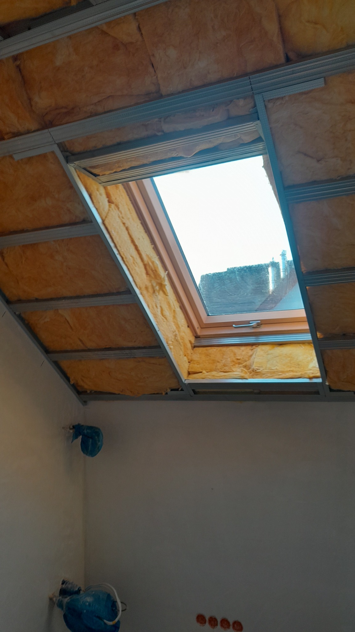 Poddasze z oknem dachowym w trakcie izolacji wełną mineralną, widoczne metalowe profile konstrukcyjne i zabezpieczone kable elektryczne.