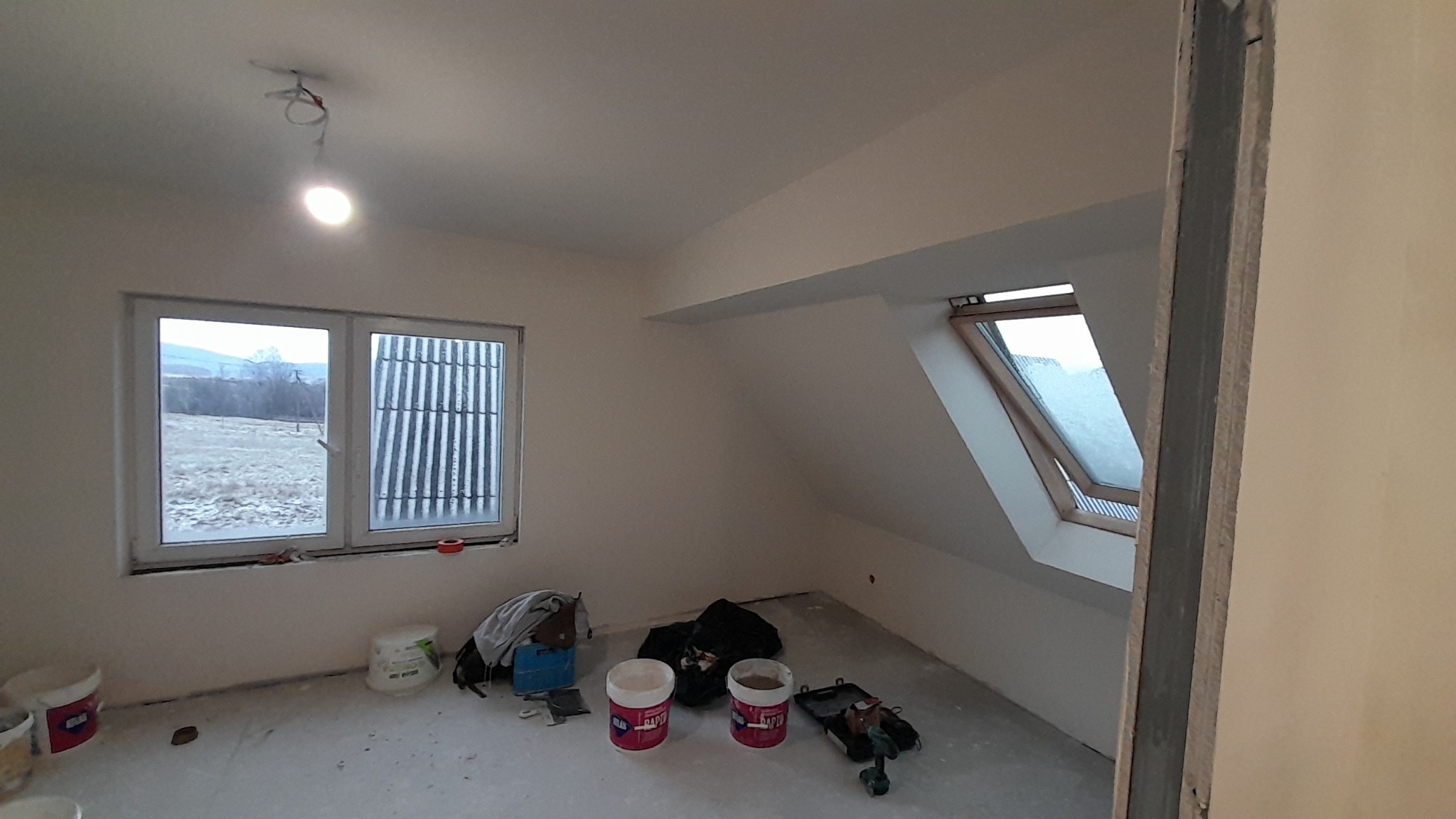Poddasze po malowaniu, z widocznymi narzędziami malarskimi na podłodze, oknem dachowym i standardowym oknem z widokiem na zimowy krajobraz za oknem.