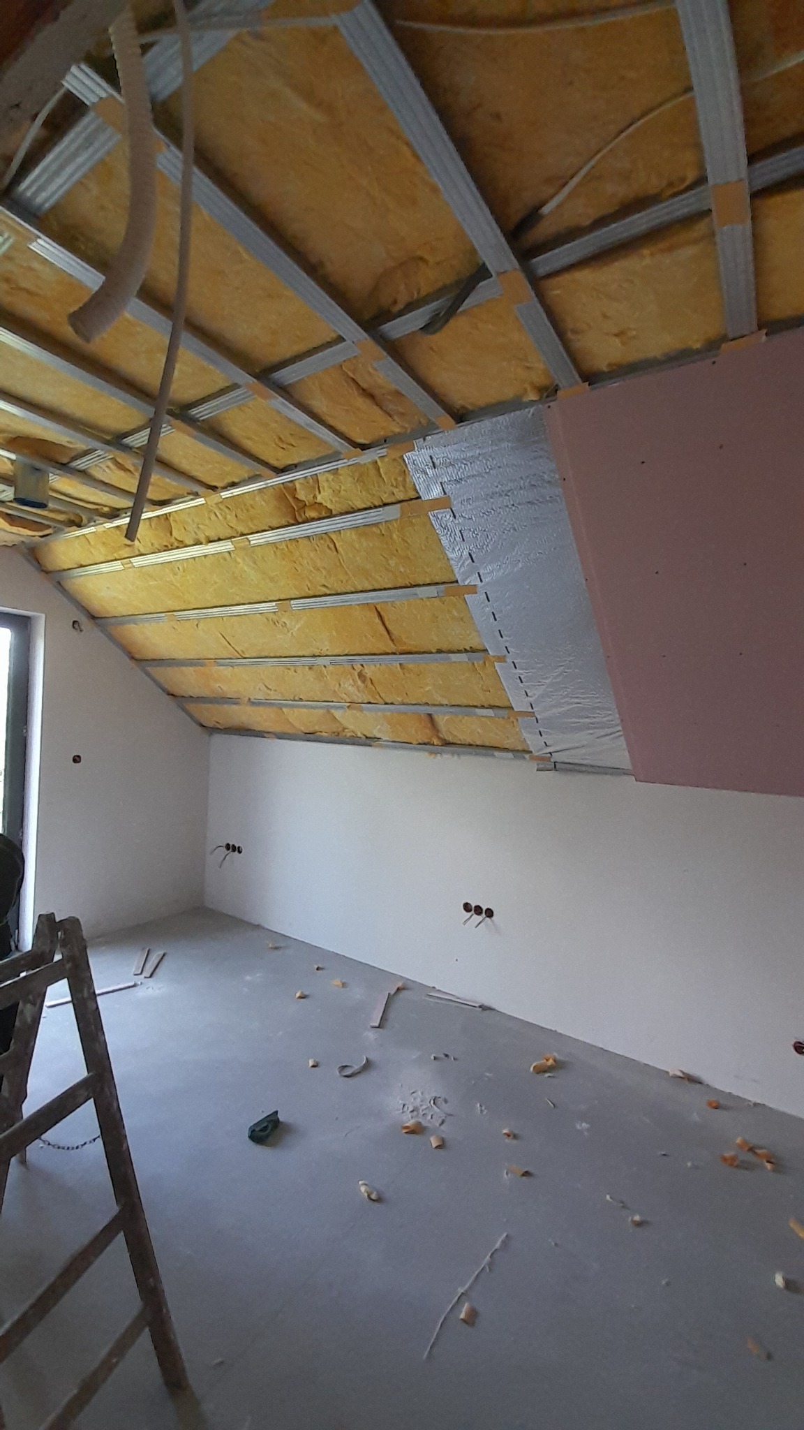 Wnętrze poddasza w trakcie prac: widoczne metalowe profile sufitowe, żółta wełna mineralna, częściowo zamontowana folia paroizolacyjna i różowa płyta gipsowo-kartonowa, drabina oparta o ścianę...