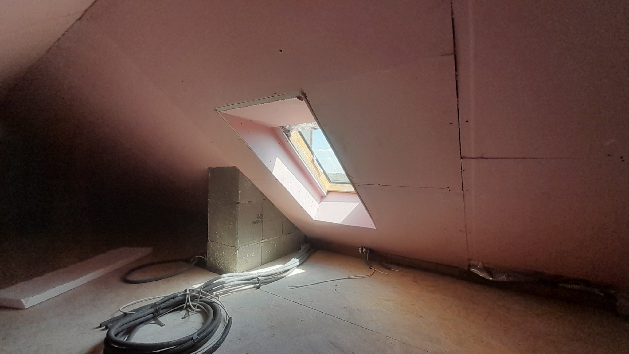 Poddasze w trakcie prac wykończeniowych: widoczne okno dachowe, ściany obłożone płytami gipsowo-kartonowymi, przewody elektryczne ułożone na podłodze, fragment izolacji.