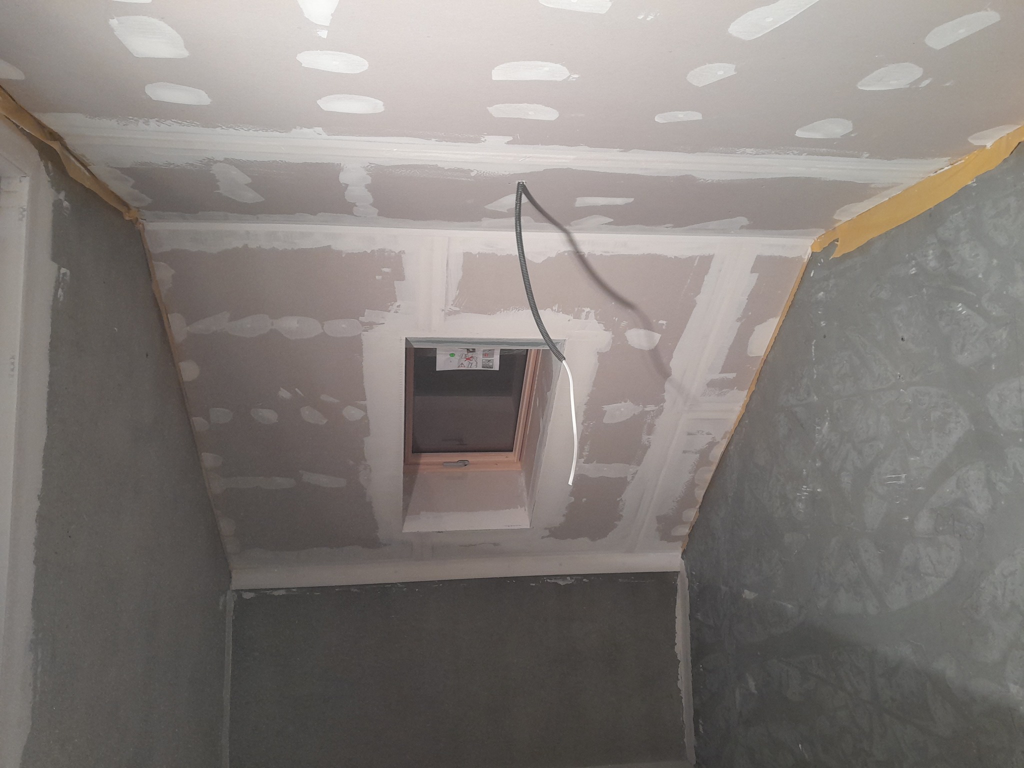 Poddasze w trakcie prac wykończeniowych: płyty karton-gips na skosach i suficie, widoczne łączenia szpachlowane, okno dachowe z drewnianą ramą, wystający kabel elektryczny. Żółta taśma malarska...