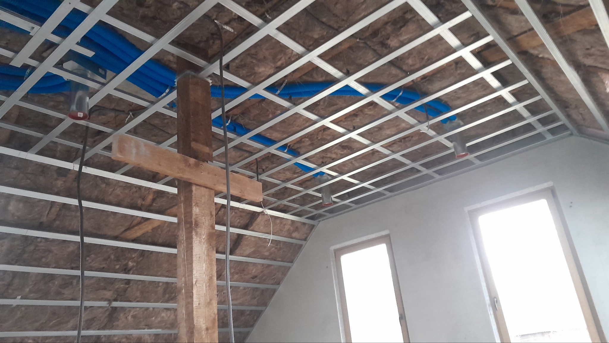 Szkielet z profili metalowych podwieszanego sufitu na poddaszu, z widoczną izolacją z wełny mineralnej i niebieskimi rurami instalacji, drewniany słup podtrzymujący konstrukcję.