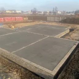 Wylany betonowy fundament z podmurówką z bloczków betonowych na działce budowlanej, w tle palety z cegłami i zabudowa jednorodzinna.