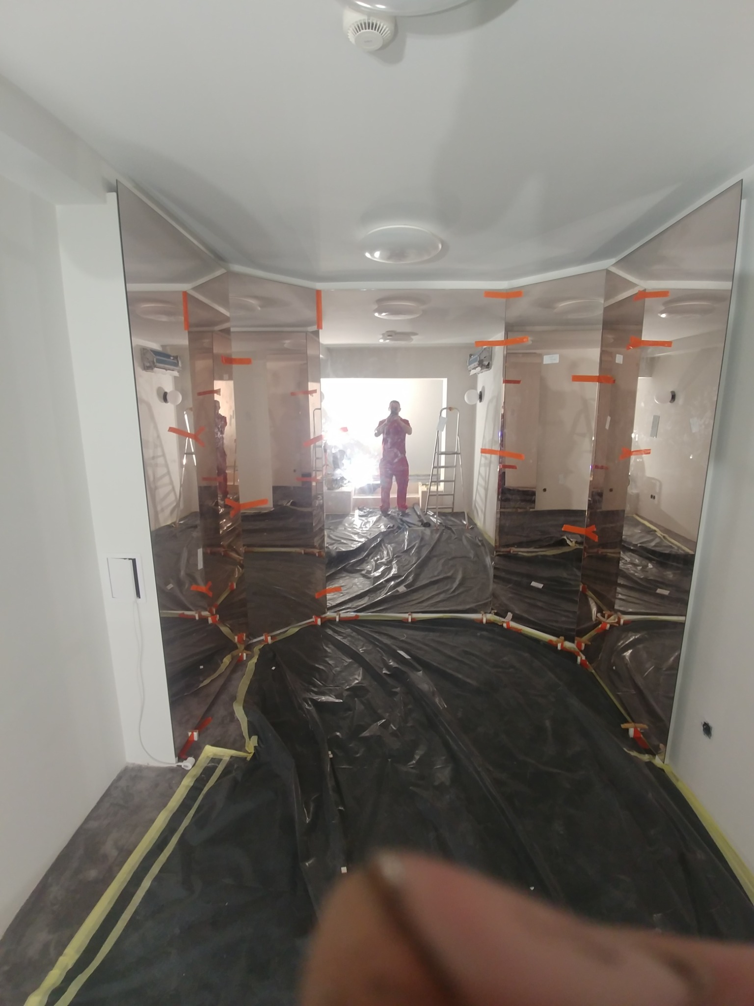 Montaż dużych, pionowych luster na ścianie, zabezpieczona podłoga folią, widoczne pomarańczowe kliny mocujące i pracownik w tle.