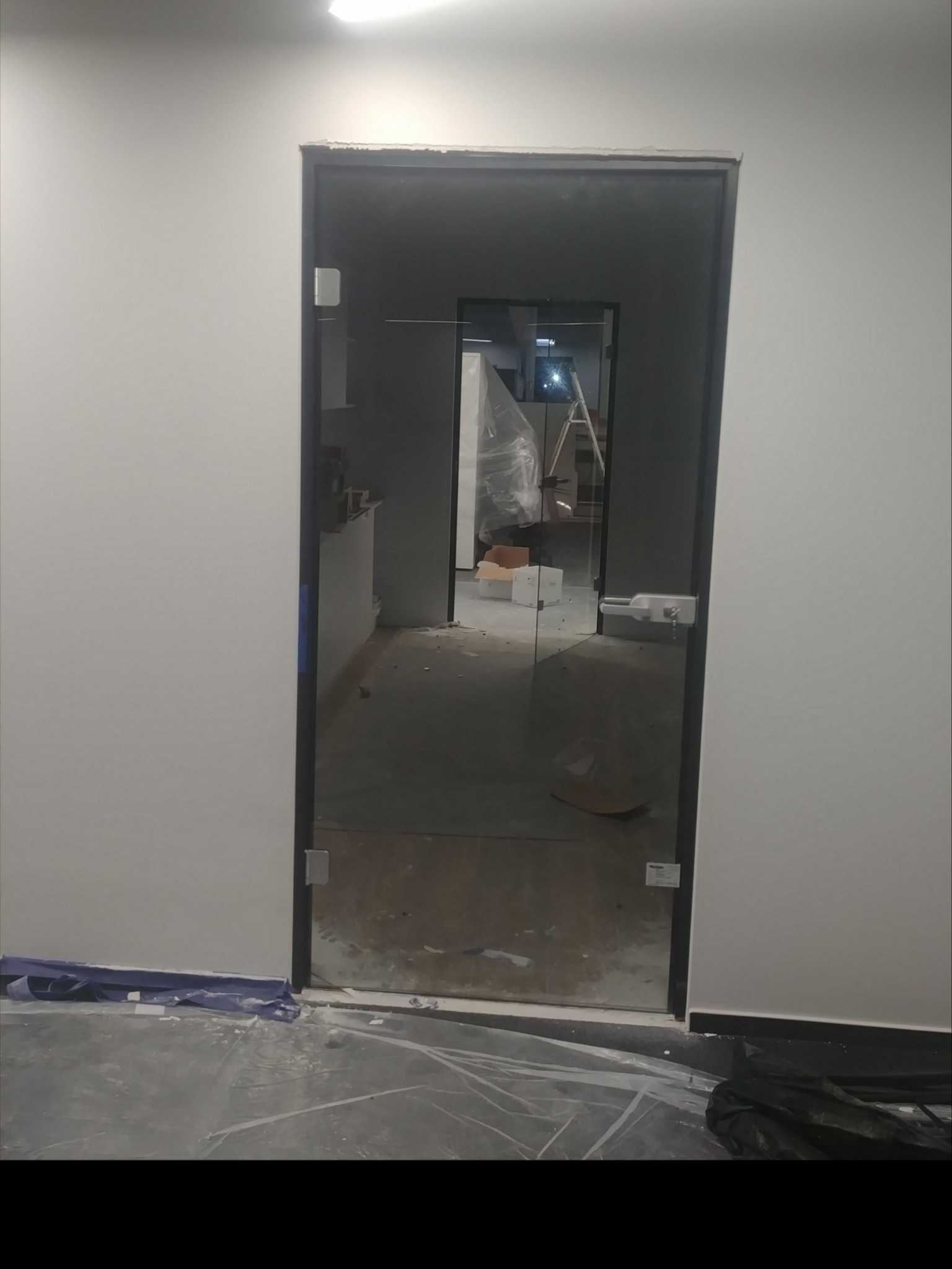 Szklane drzwi z czarną ramą zamontowane w biurze w Warszawie, widoczne zabezpieczenia podłogi folią malarską, w tle drabina i materiały budowlane.