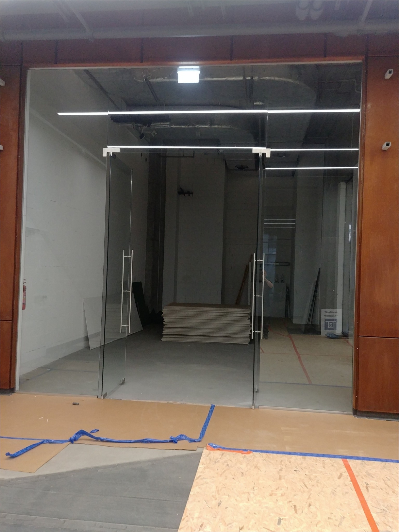 Szklane drzwi wejściowe z metalowymi uchwytami, zabezpieczone kartonem i taśmą na podłodze w surowym wnętrzu budynku.