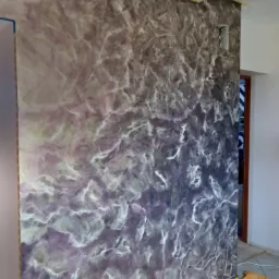 Ściana z dekoracyjną, fakturowaną powierzchnią w odcieniach fioletu, szarości i bieli, zabezpieczona żółtą taśmą malarską przy podłodze i suficie, z widocznym gniazdkiem elektrycznym w trakcie...