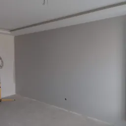 Świeżo pomalowane szare ściany w pokoju, widoczna lampa budowlana na statywie i wystające kable z sufitu.