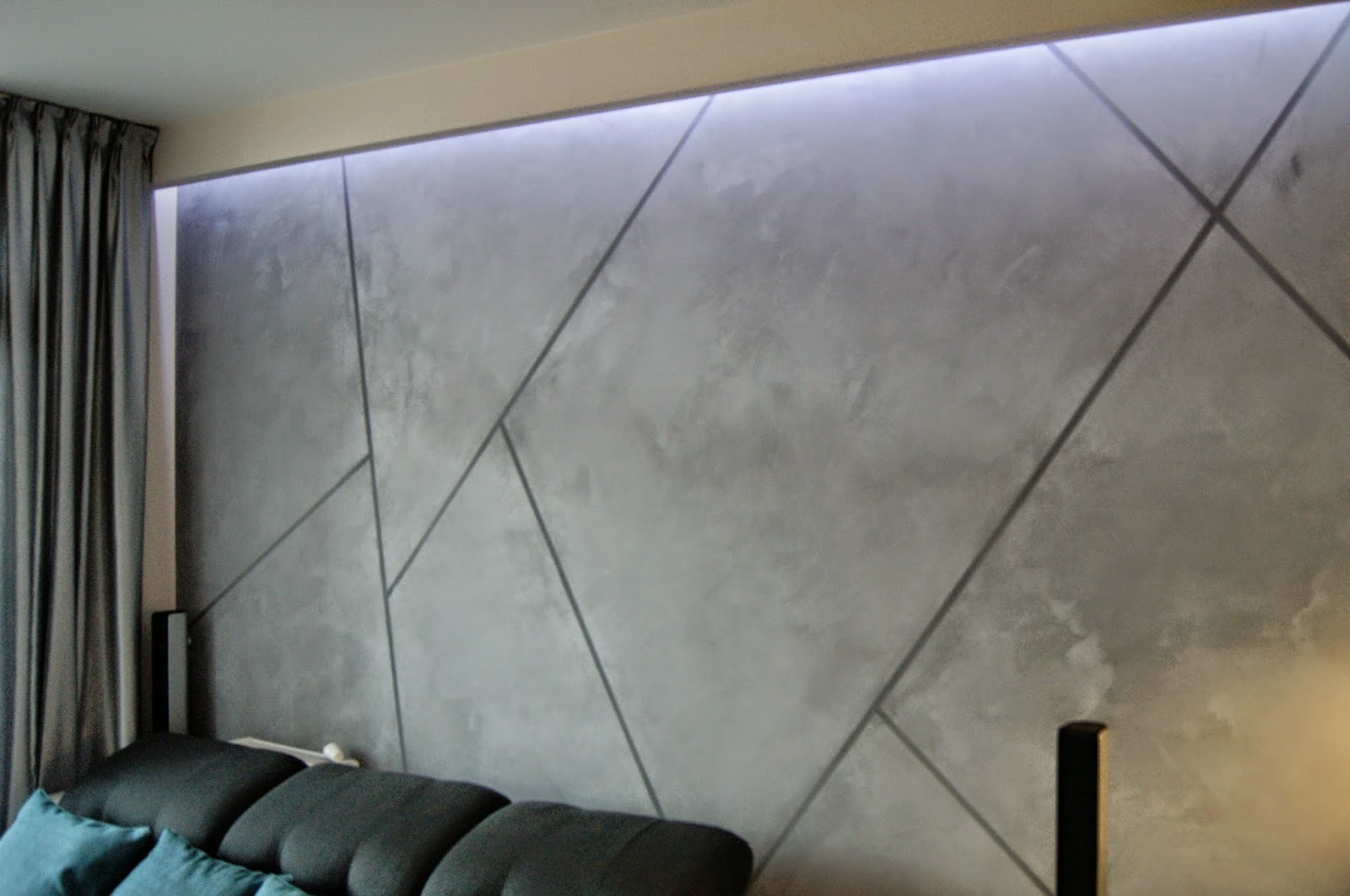 Ściana w salonie wykończona dekoracyjną szarą gładzią z geometrycznym wzorem podkreślonym cienkimi, ciemnymi liniami, oświetlona listwą LED podsufitową, widoczny fragment zasłony i sofy.