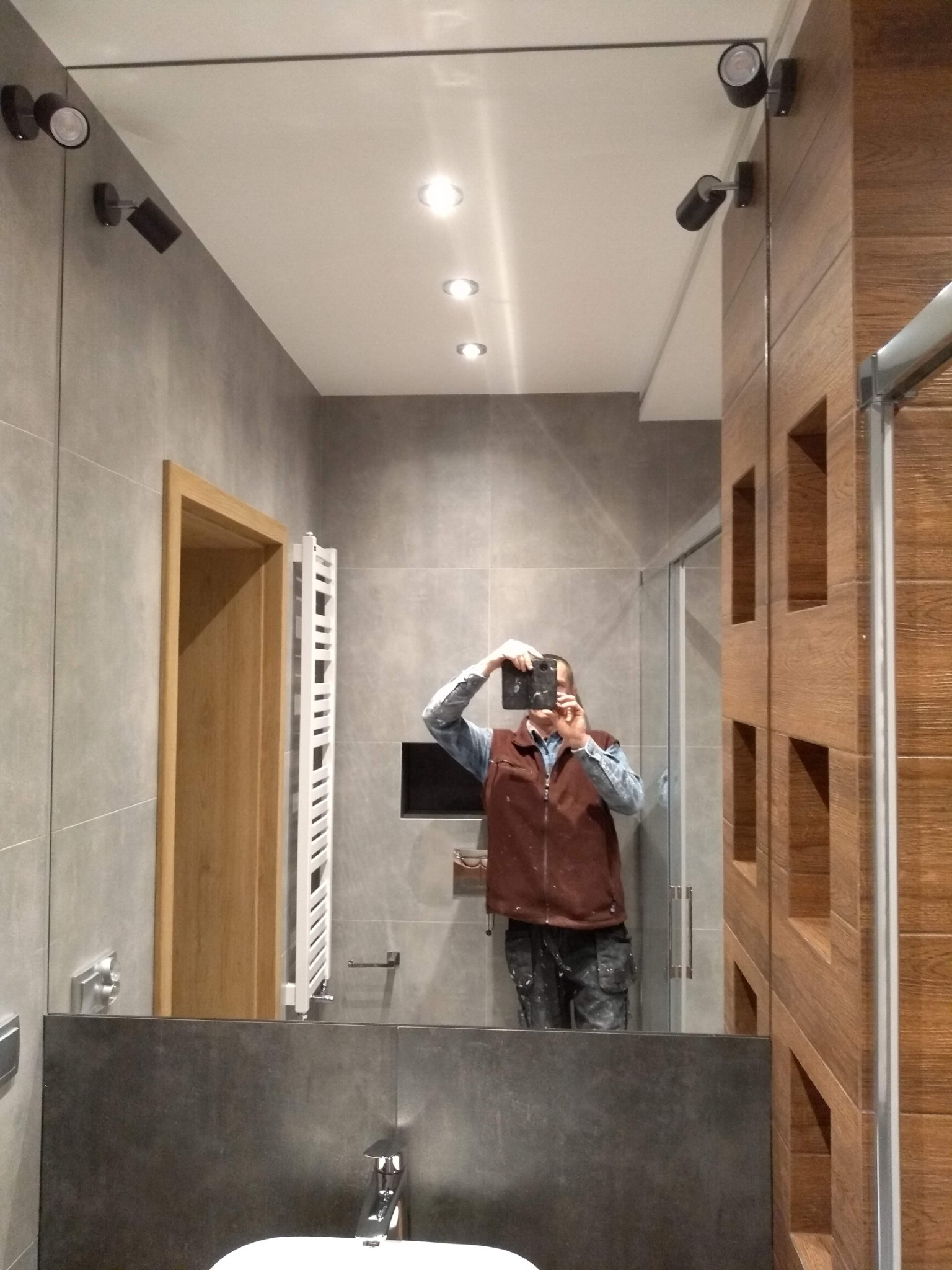 Nowoczesna łazienka z szarymi płytkami, drewnianymi akcentami i oświetleniem punktowym, odbicie osoby robiącej zdjęcie w lustrze.