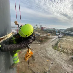 Pracownik w kasku i uprzęży, zwisający na linach z pionowej, ciemnej konstrukcji przemysłowej, z widokiem na port, autostradę i pochmurne niebo.