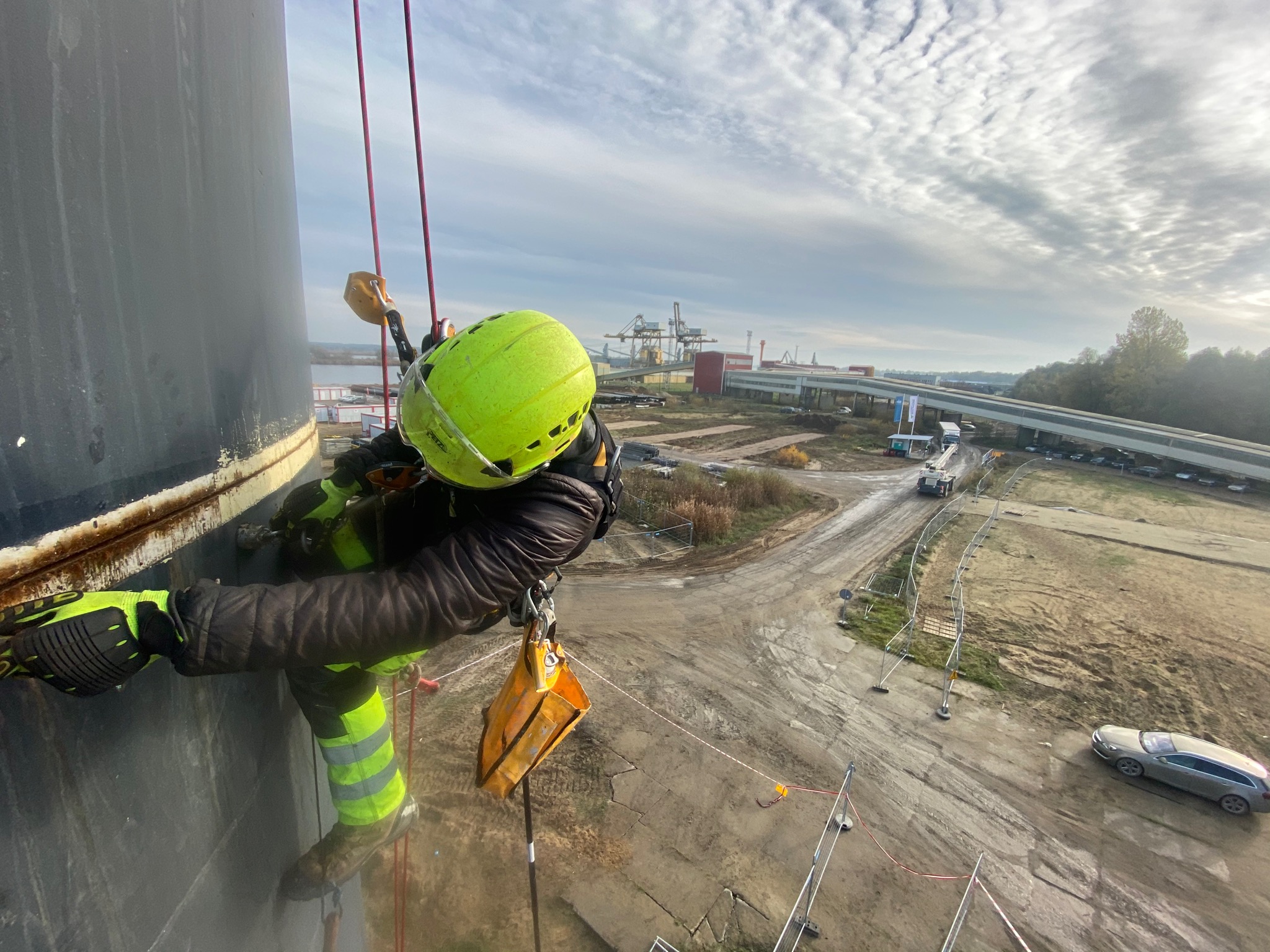 Pracownik w kasku i uprzęży, zwisający na linach z pionowej, ciemnej konstrukcji przemysłowej, z widokiem na port, autostradę i pochmurne niebo.