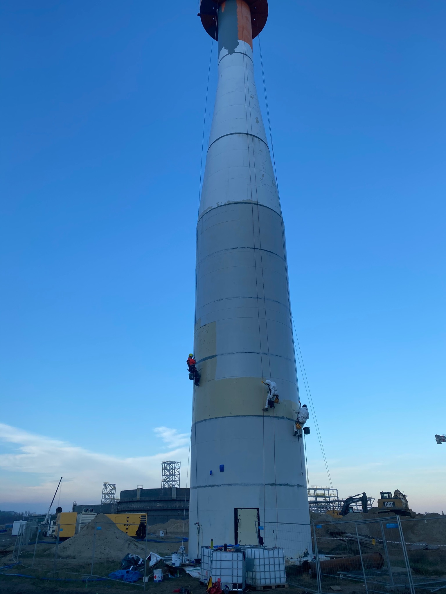 Dwóch pracowników na linach zabezpieczających, w kombinezonach, wykonuje prace na wysokościach na dużej, cylindrycznej konstrukcji. Jeden robotnik nakłada szpachlę, a drugi maluje. W tle błękitne...