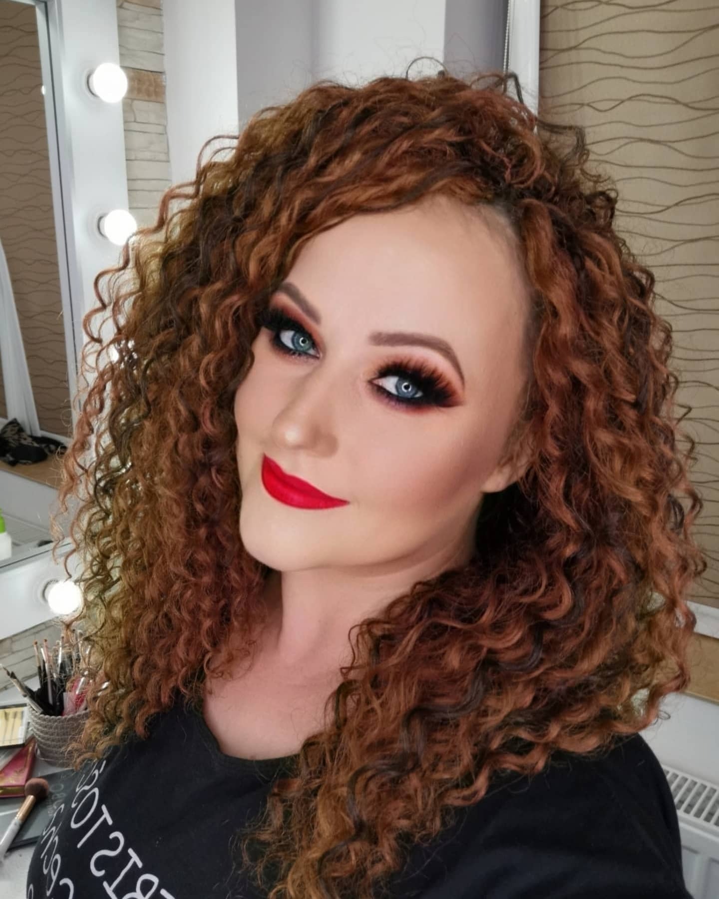 Portret kobiety z intensywnym makijażem oczu i czerwonymi ustami, burza rudych loków, widoczne oświetlenie studyjne.