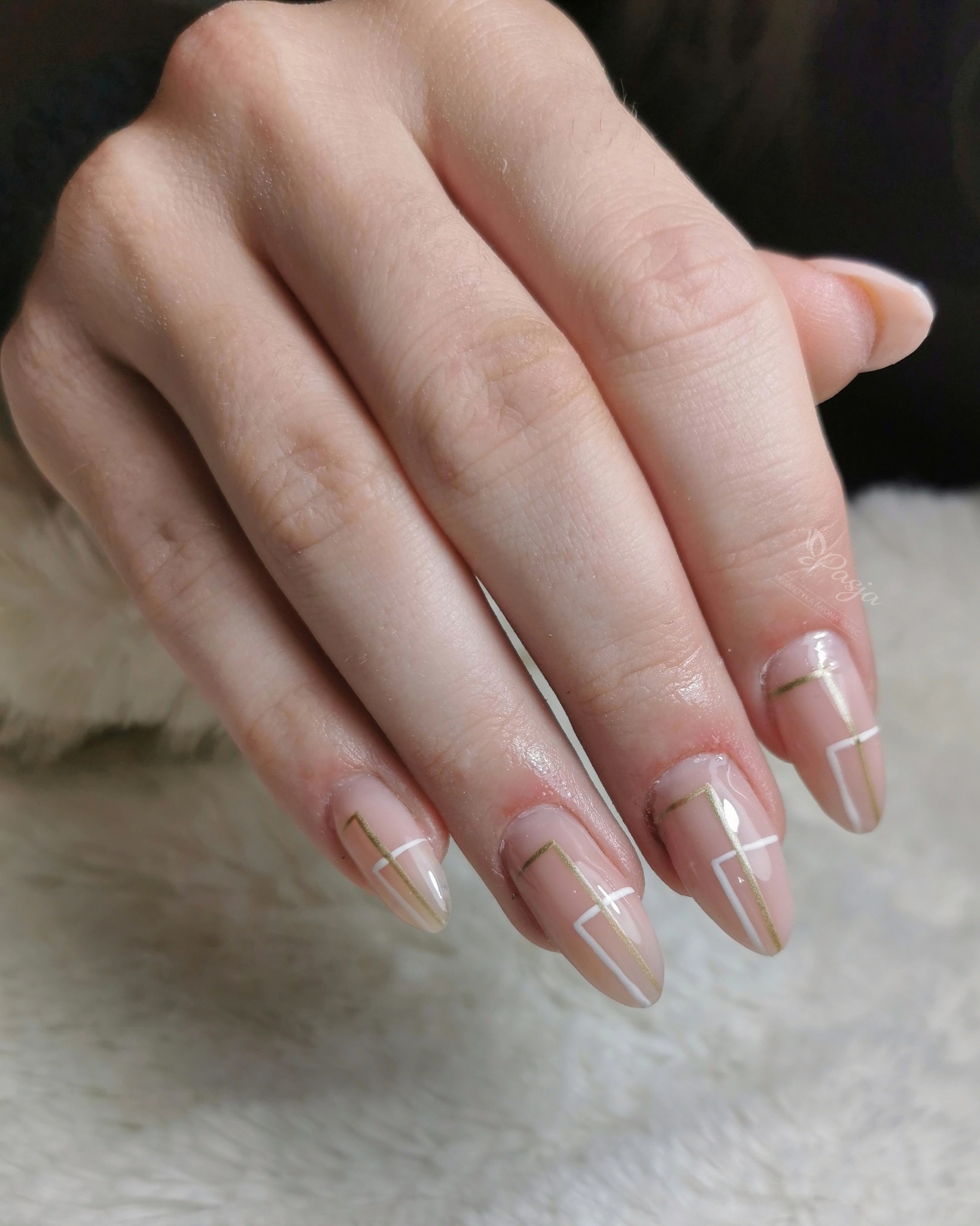 Dłoń z jasnym, błyszczącym manicure hybrydowym z geometrycznym wzorem w kolorze białym i złotym na beżowym tle.