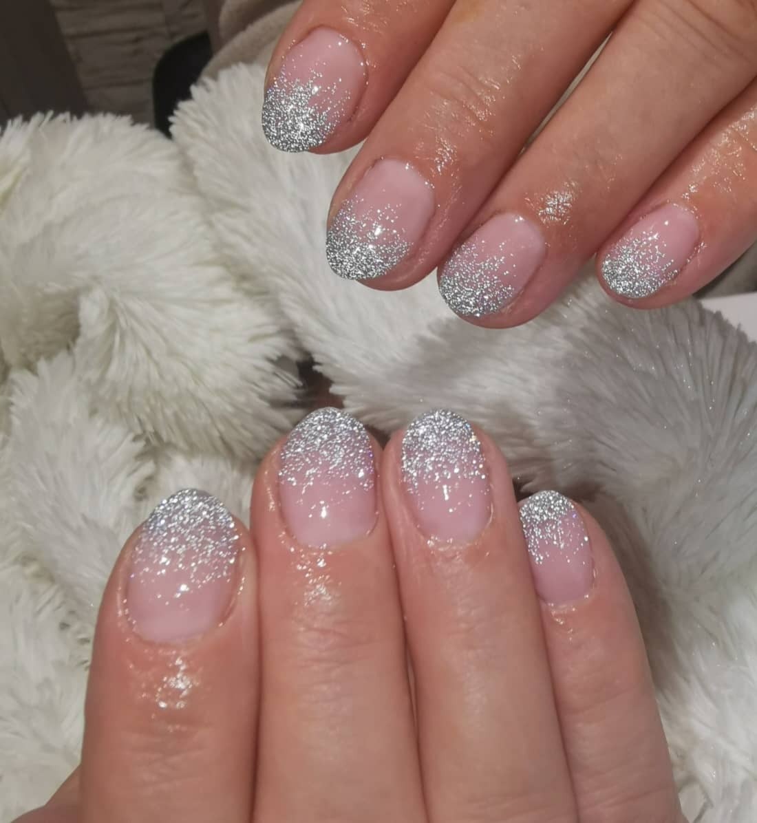 Dłonie z świeżo wykonanym manicure hybrydowym z efektem srebrnego brokatu na końcówkach, ułożone na puszystym białym tle.