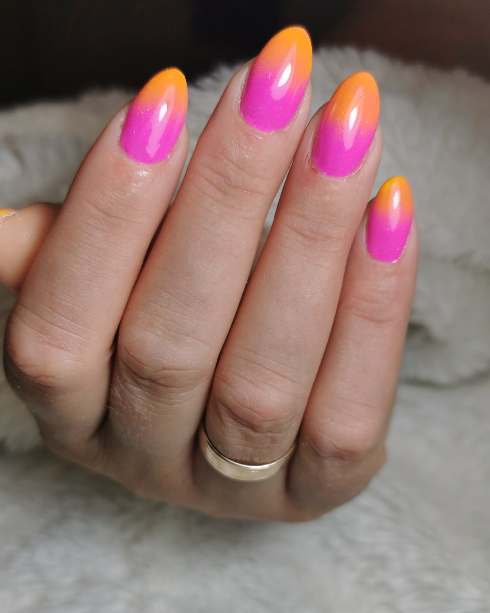 Dłoń z owalnymi paznokciami pomalowanymi lakierem w odcieniach różu i pomarańczy, tworząc efekt ombre; na palcu serdecznym złoty pierścionek na tle białego, puszystego materiału.