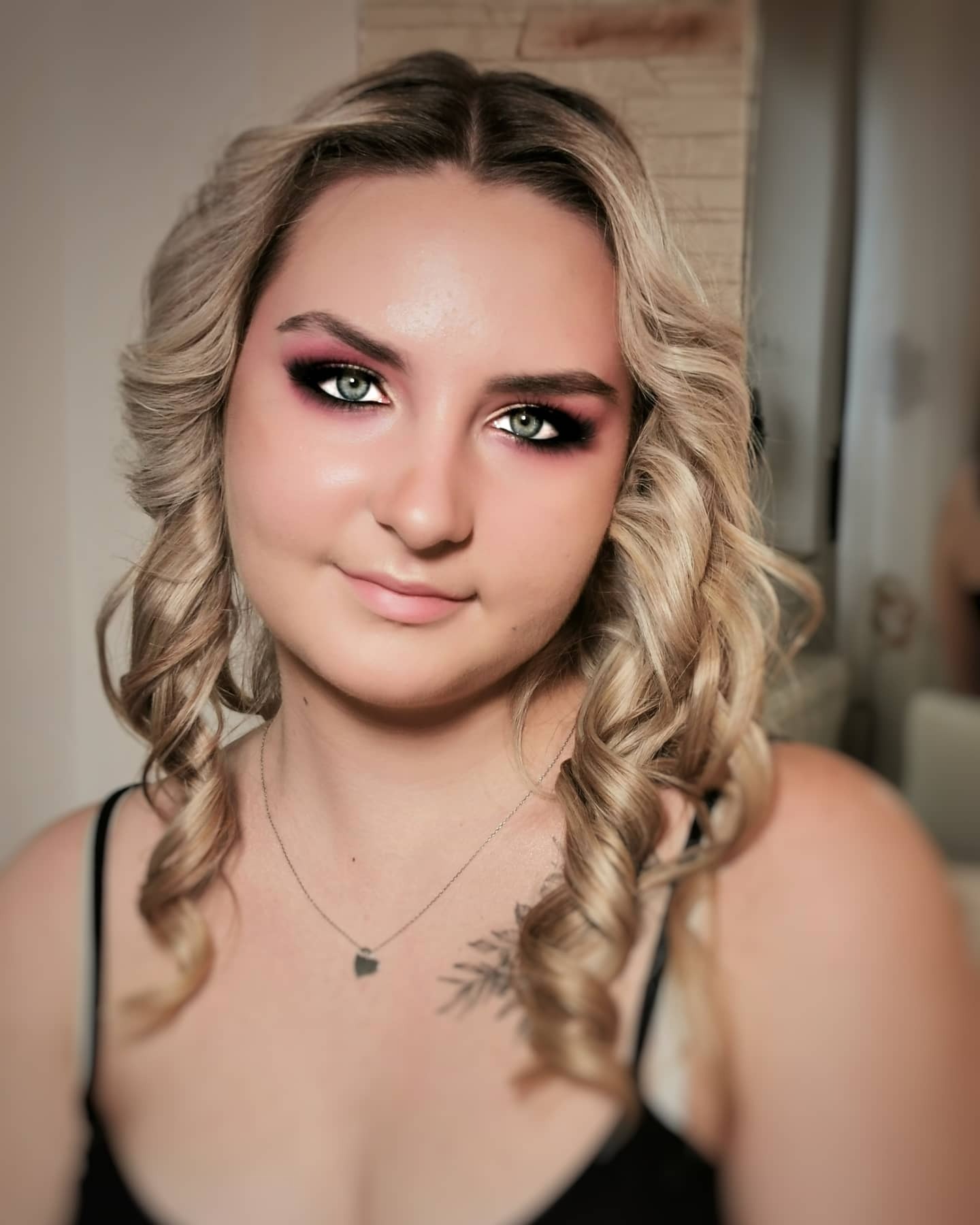 Portret blondynki z intensywnym, różowo-czarnym cieniowaniem powiek i delikatnym uśmiechem, podkreślonym tatuażem roślinnym na dekolcie i srebrnym naszyjnikiem.