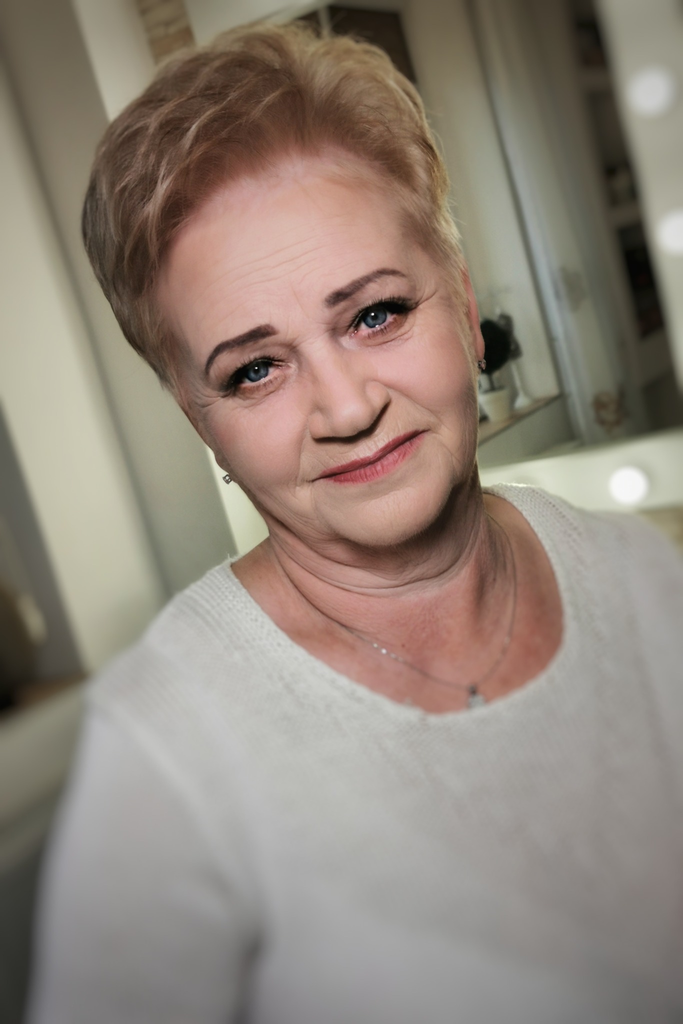 Portret uśmiechniętej kobiety z delikatnym makijażem, krótkimi blond włosami i srebrnym naszyjnikiem, uchwycony z bliska.