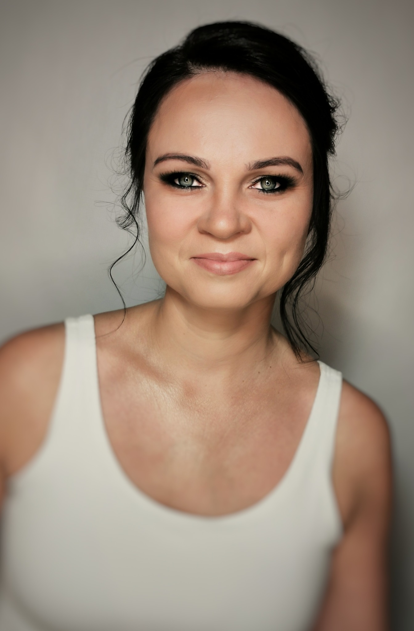 Portret kobiety z ciemnymi włosami upiętymi w kok, z wyraźnie podkreślonymi oczami i delikatnym uśmiechem, na jasnym tle.