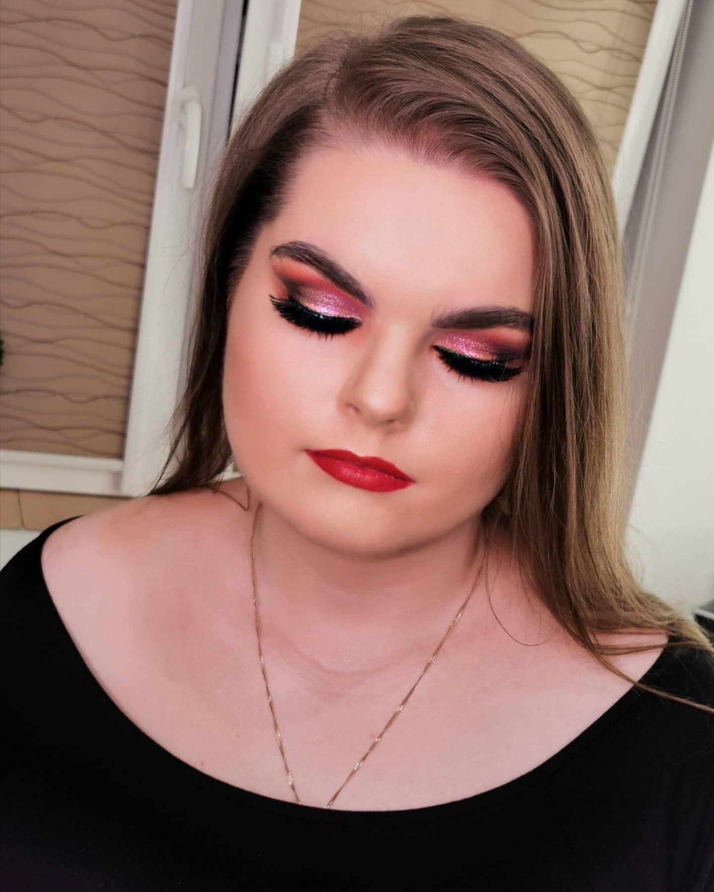 Portret kobiety z intensywnym, błyszczącym makijażem oczu w odcieniach różu i czerwieni, oraz wyrazistymi czerwonymi ustami, patrzącej w dół, z długimi, prostymi włosami i delikatnym złotym...