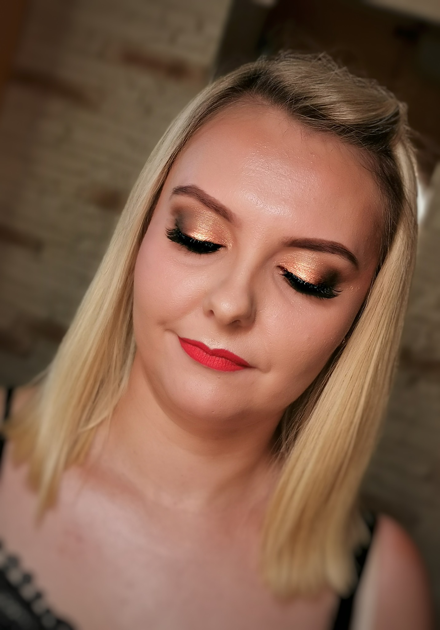 Portret kobiety z blond włosami i profesjonalnym makijażem: złote cienie z brokatem i sztuczne rzęsy, matowa czerwona szminka, delikatny kontur twarzy.