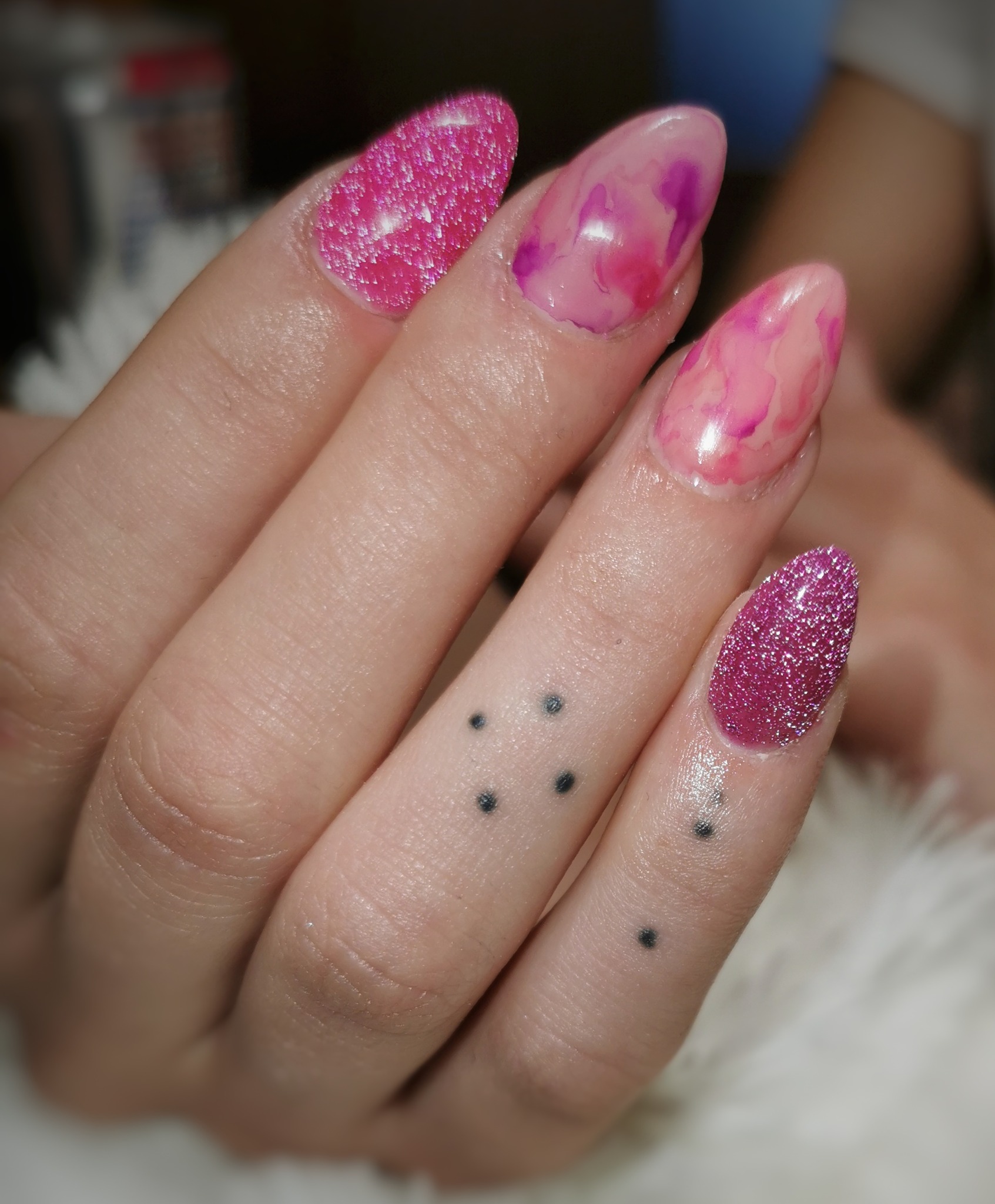 Zbliżenie dłoni z pomalowanymi paznokciami: dwa brokatowe w odcieniach różu, dwa z efektem marmuru w różach i fioletach; na palcu tatuaż z sześciu kropek.