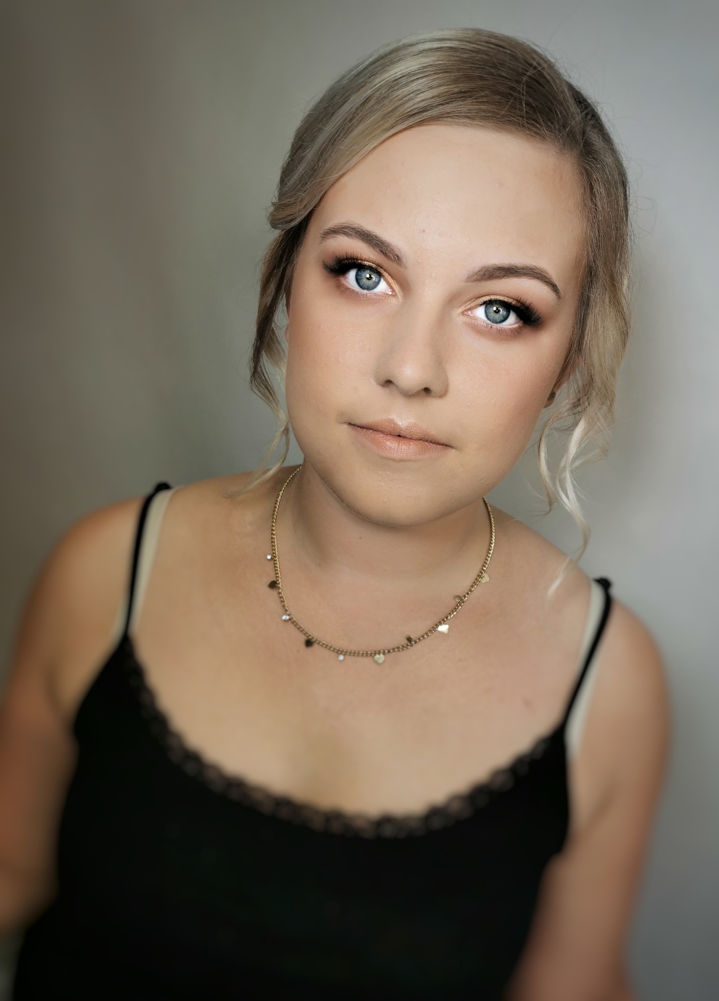 Portret kobiety z subtelnym makijażem oczu i ust, włosy upięte z przodu, delikatny złoty naszyjnik z zawieszkami w kształcie serc, czarny top na ramiączkach.
