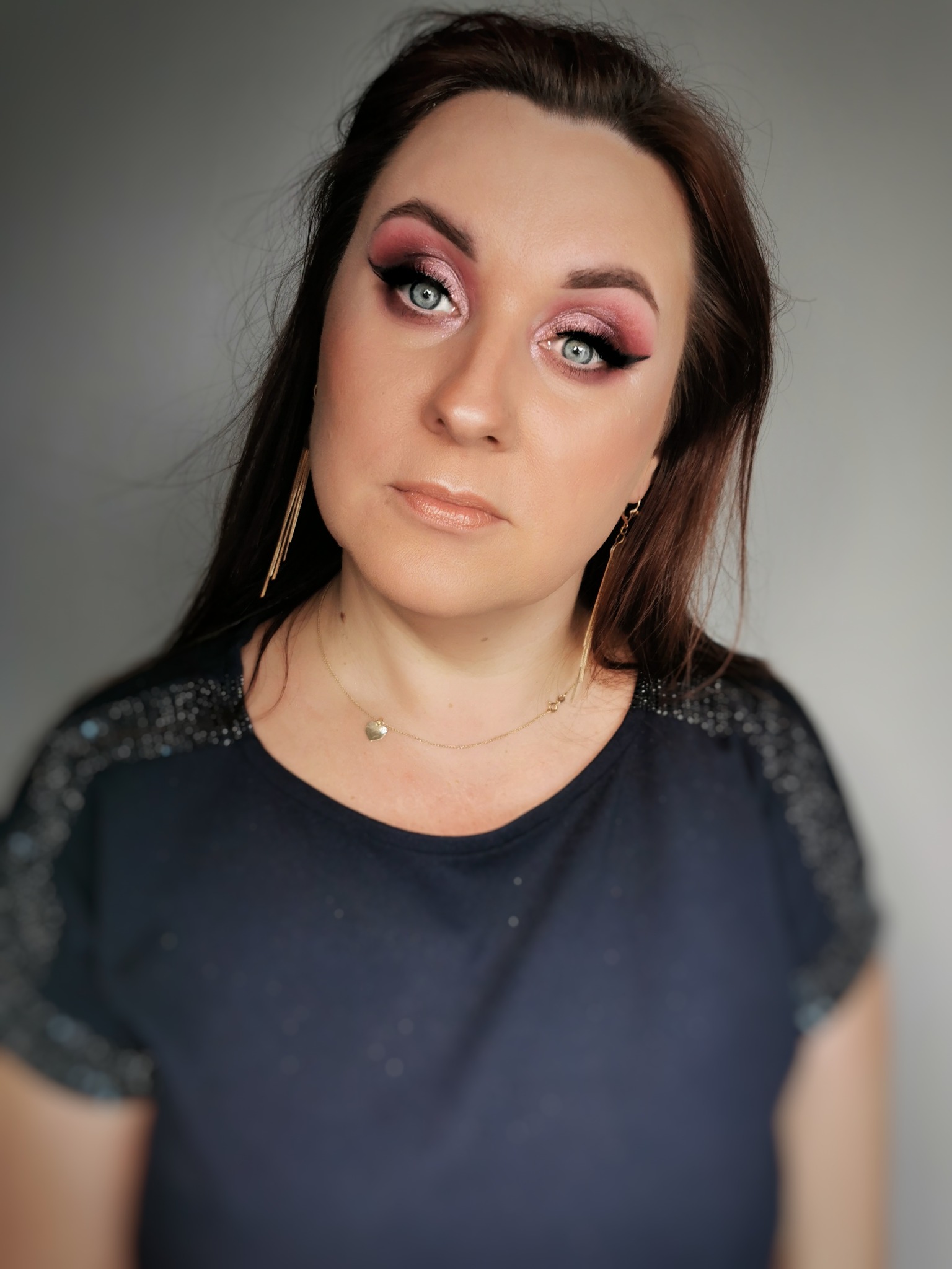 Portret kobiety z intensywnym różowym makijażem oczu, podkreślonym czarnym eyelinerem, długimi złotymi kolczykami i delikatnym błyszczykiem na ustach, na szarym tle.