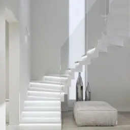 Minimalistyczne wnętrze z białymi schodami podświetlonymi ledami, szklanymi balustradami, pufą i srebrnymi wazonami na drewnianej podłodze.