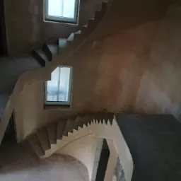 Wnętrze w stanie surowym z widocznymi schodami i świeżo nałożonymi tynkami maszynowymi, okna zabezpieczone folią, widok z góry.