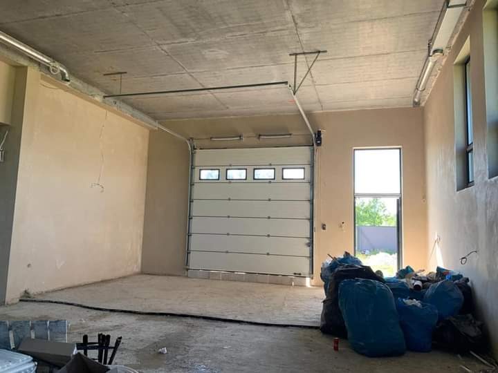 Wnętrze surowego pomieszczenia z niedokończonymi ścianami i częściowo otwartą segmentową bramą garażową z oknami, worki z odpadami budowlanymi w rogu, widoczne instalacje pod sufitem.