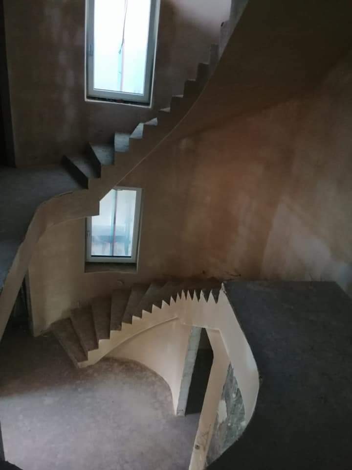 Wnętrze w stanie surowym z widocznymi schodami i świeżo nałożonymi tynkami maszynowymi, okna zabezpieczone folią, widok z góry.