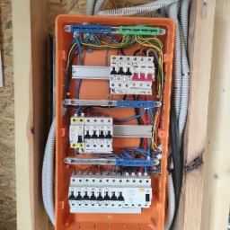 Pomarańczowa skrzynka rozdzielcza z bezpiecznikami i przewodami, zamontowana na drewnianej ścianie z widocznymi rurami osłonowymi.