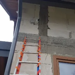 Kamera monitoringu zamontowana na szarej ścianie budynku z pustaków, widoczna drabina aluminiowa z pomarańczowymi elementami zabezpieczającymi oraz rura spustowa