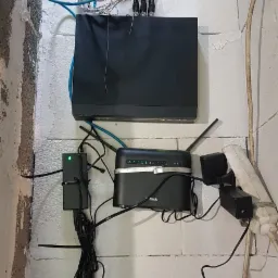 Rejestrator monitoringu, router D-Link i zasilacze zamontowane na surowej, pomalowanej na biało ścianie z betonu komórkowego. Widoczne liczne przewody połączeniowe.