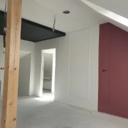 Wnętrze w trakcie prac wykończeniowych: surowa drewniana belka kontrastuje z pomalowaną na biało ścianą z dekoracyjną ramą i bordową ścianą z panelem, widoczne przygotowania do montażu oświetlenia...