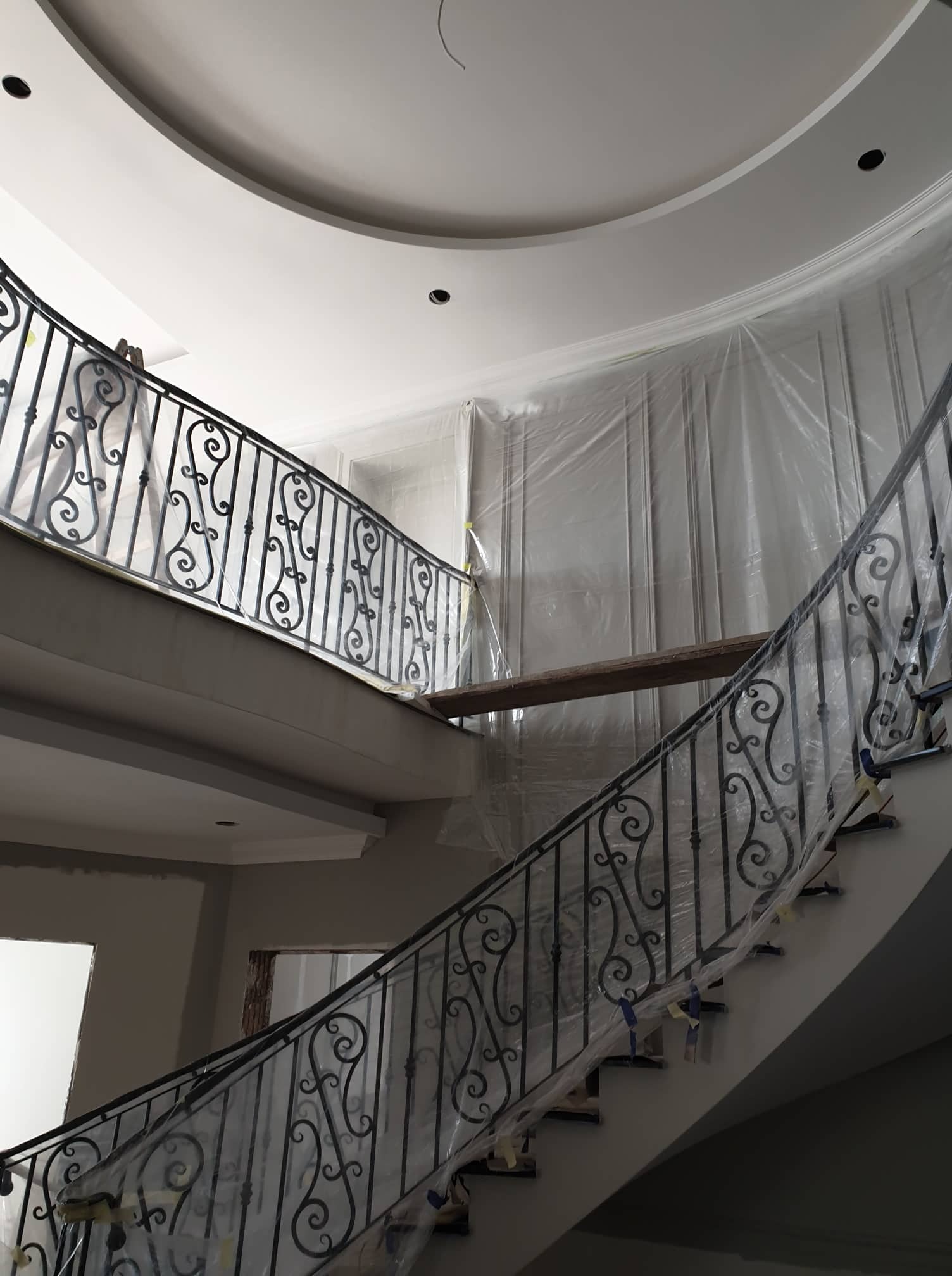Wnętrze w trakcie remontu: kręte schody z ozdobną, metalową balustradą zabezpieczoną folią malarską, ściany z panelami dekoracyjnymi również zakryte folią, sufit z okrągłą wnęką.